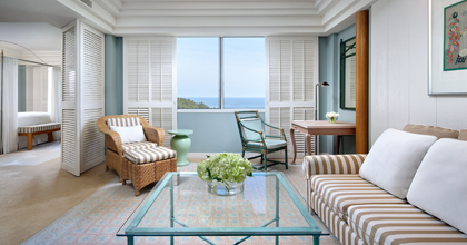 Image of a corner suite living room with pastel-toned interiors, a sofa for two, a table, rattan chairs, and a dining table overlooking the sea.